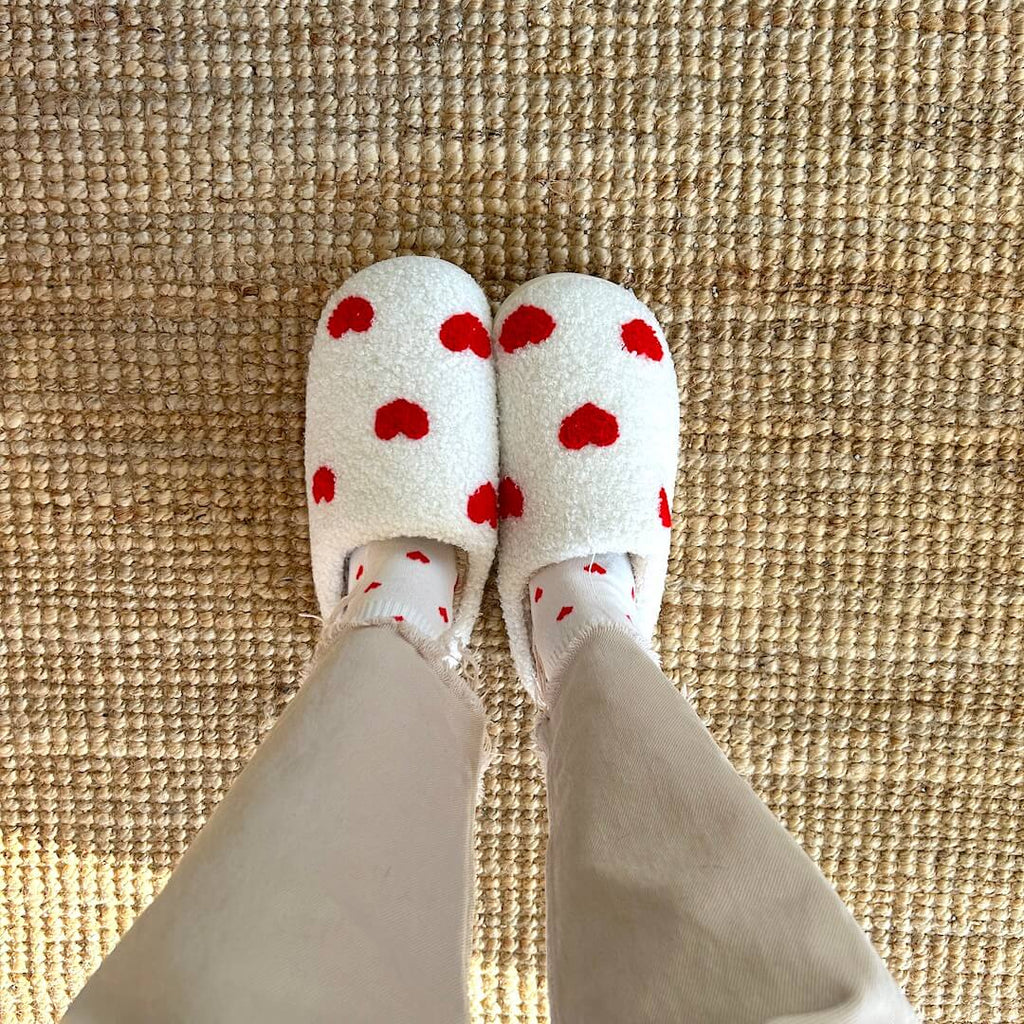 Person wearing heart-patterned slippers and socks on a rug.