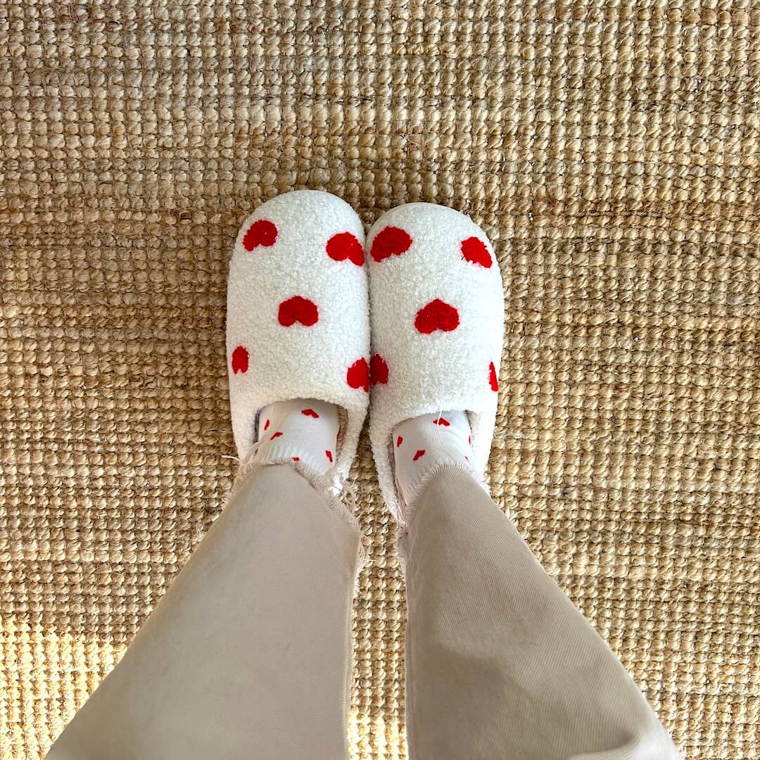 Person wearing heart-patterned slippers and socks on a rug.