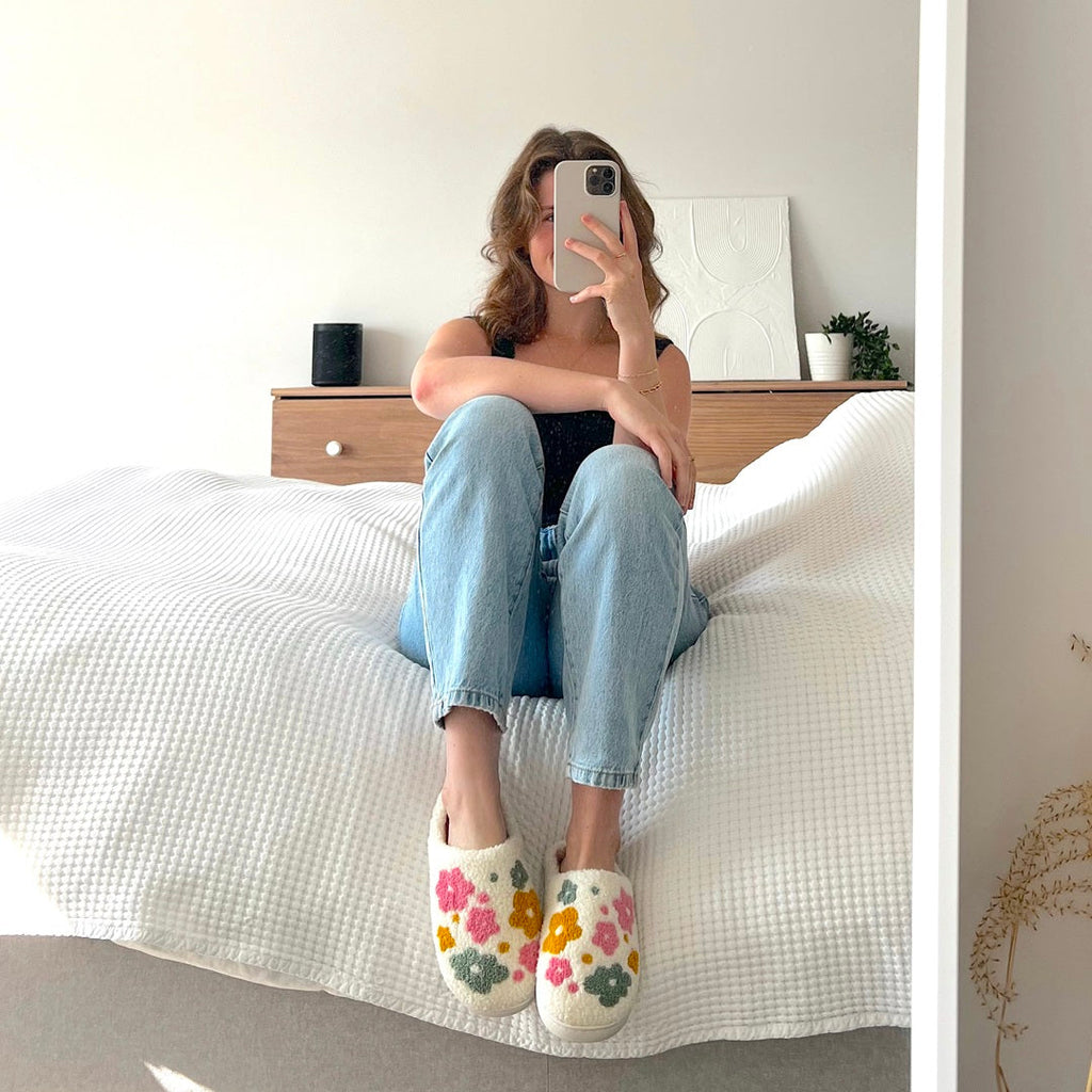 Woman in jeans and floral slippers sits on a bed.