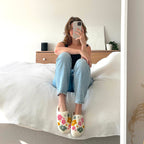 Woman in jeans and floral slippers sits on a bed.